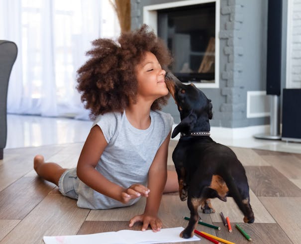 Jente ligger på gulv, en hund snuser henne i ansiktet.